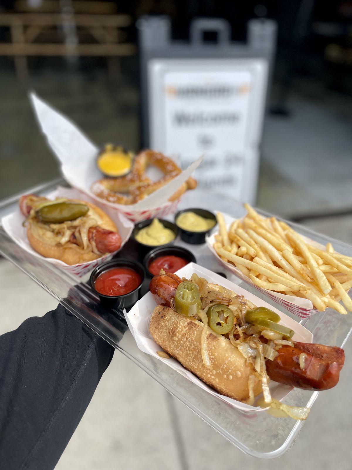 A tray held out at an event with two sausages on rolls topped with grilled onions and jalapeños, a soft pretzel with cheese sauce, fries, and cups of ketchup and mustard