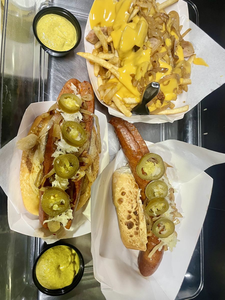 Overhead view of two sausages on rolls topped with sauerkraut, grilled onions and jalapeño slices, next to cheese fries with grilled onions and two cups of mustard