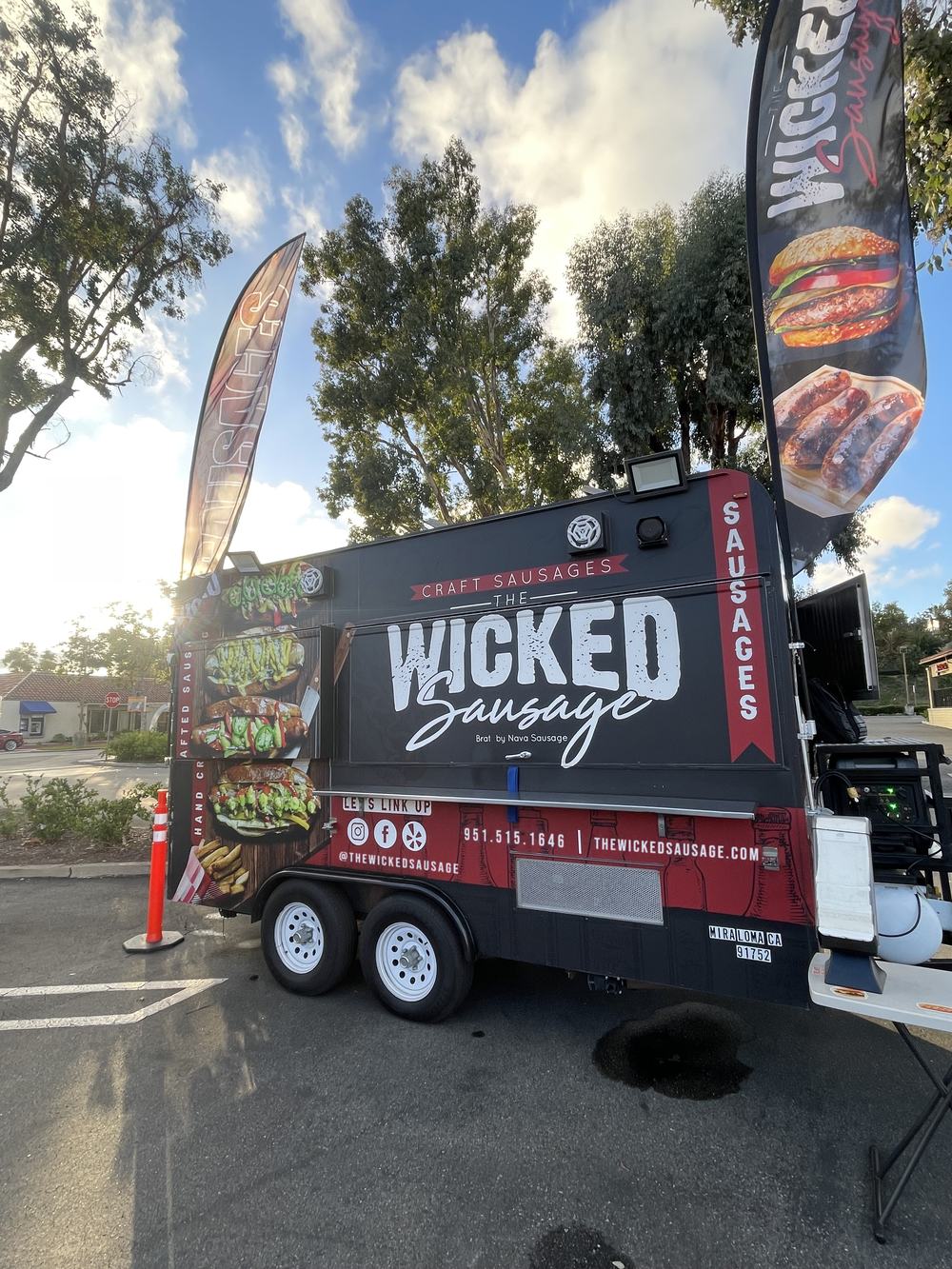 The Wicked Sausage trailer parked in a lot under a blue sky: a matte black trailer with red accents, white WICKED Sausage lettering, sausage sandwich graphics on the side, and tall feather flags on either side